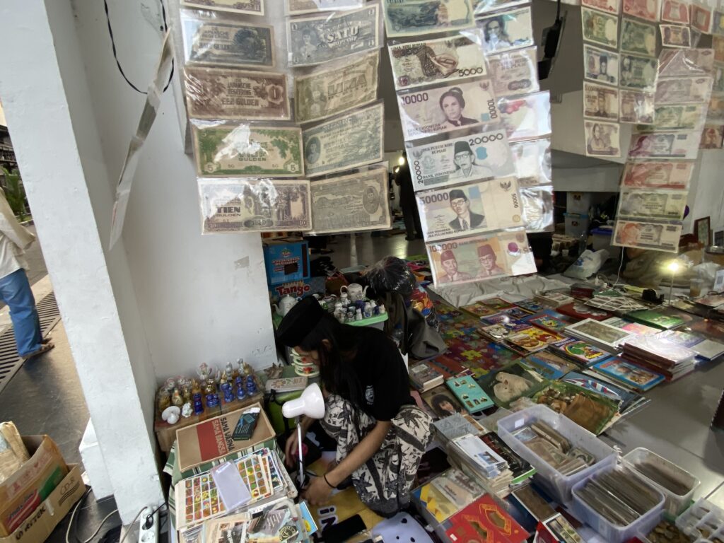 Store interior with rows of laminated currency notes hanging from strings; a person sits at a cluttered counter handling papers and small items.