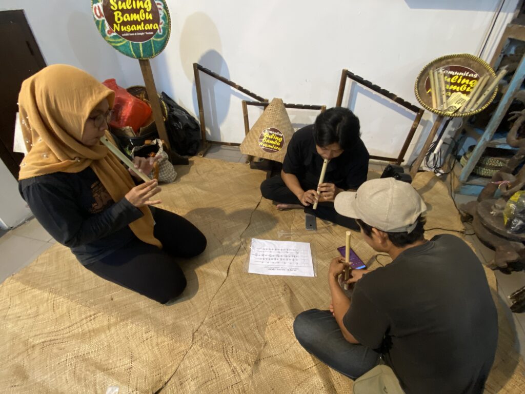 Group of people seated on a woven mat indoors, playing bamboo flutes with a music sheet on the floor.