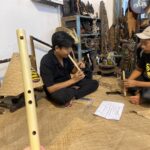 Two men sit on a woven mat, one playing a long bamboo flute while the other holds a shorter instrument, in a craft shop filled with carvings and shelves of objects.