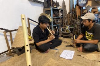 Two men sit on a woven mat, one playing a long bamboo flute while the other holds a shorter instrument, in a craft shop filled with carvings and shelves of objects.