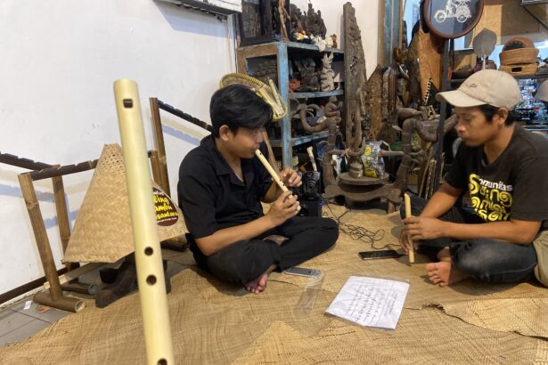 Two men sit on a woven mat, one playing a long bamboo flute while the other holds a shorter instrument, in a craft shop filled with carvings and shelves of objects.