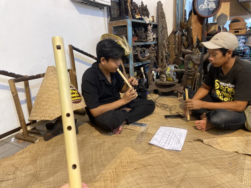 Two men sit on a woven mat, one playing a long bamboo flute while the other holds a shorter instrument, in a craft shop filled with carvings and shelves of objects.