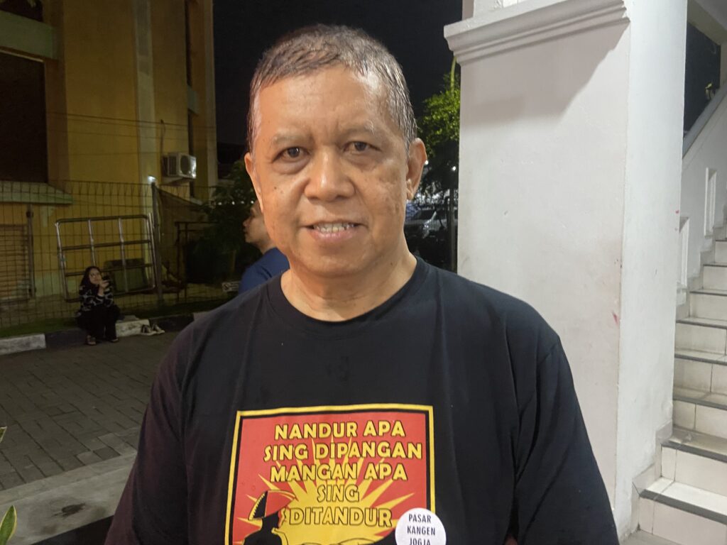 Close-up of a middle-aged man with short gray hair wearing a black T-shirt with a red-yellow graphic, standing outdoors at night beside a white column.