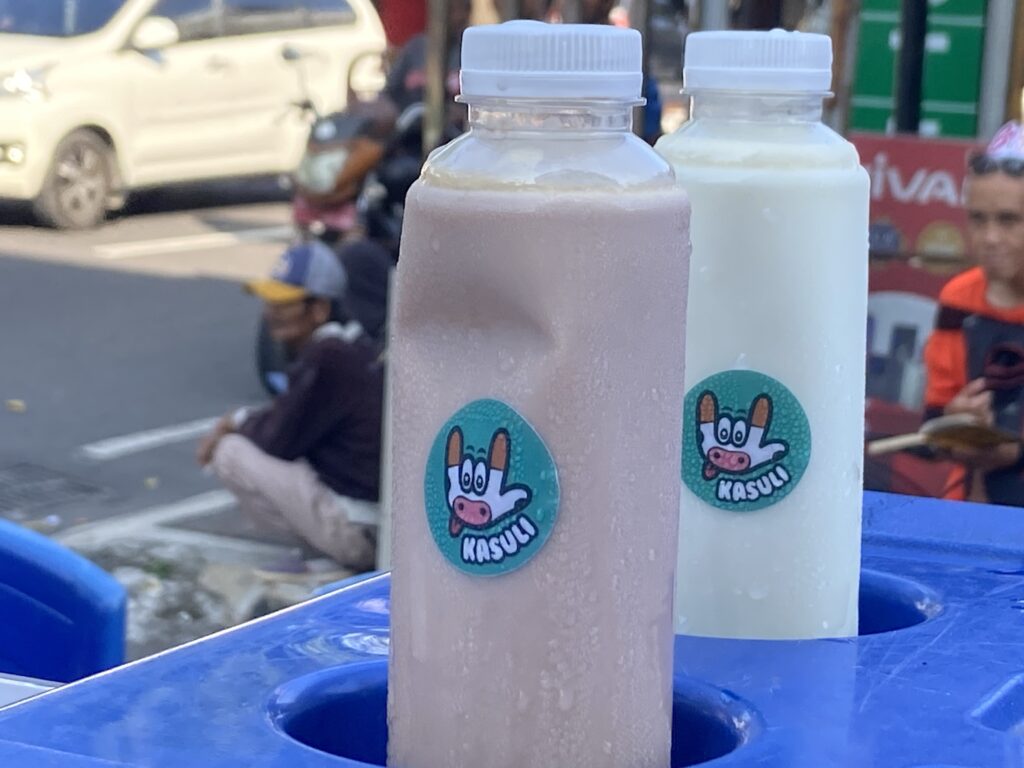 IMG_9345 - Mabur.co Two squeeze bottles with white caps on a blue table in a street scene; each bottle has a round sticker with a cartoon cow face and the word Kasuli (partial).