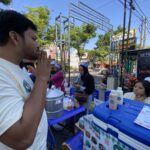 Side-profile of a man in a light shirt at an outdoor street market, standing by a vendor table with blue coolers and menus visible in the foreground