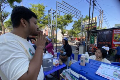 Side-profile of a man in a light shirt at an outdoor street market, standing by a vendor table with blue coolers and menus visible in the foreground