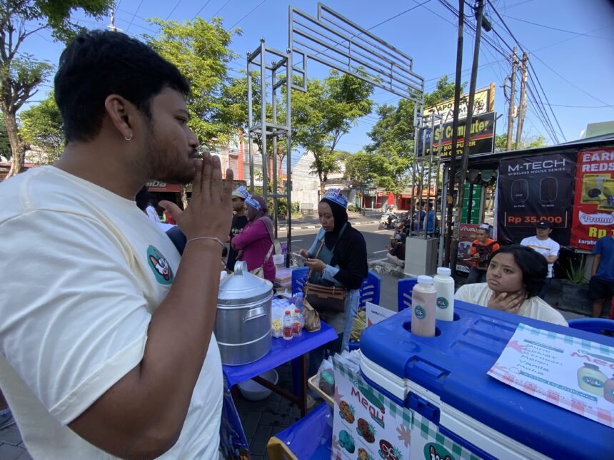 IMG_9347 1 - Mabur.co Side-profile of a man in a light shirt at an outdoor street market, standing by a vendor table with blue coolers and menus visible in the foreground