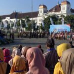 Large crowd in colorful traditional clothing watching a ceremony in a city square, with a brass band on steps and a large blue event banner in front of a white historic building with copper domes.