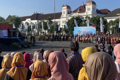 Large crowd in colorful traditional clothing watching a ceremony in a city square, with a brass band on steps and a large blue event banner in front of a white historic building with copper domes.