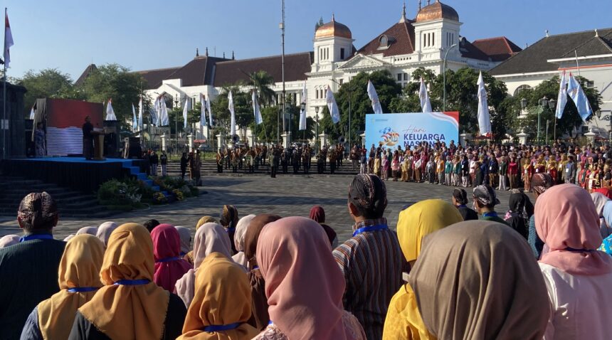 Large crowd in colorful traditional clothing watching a ceremony in a city square, with a brass band on steps and a large blue event banner in front of a white historic building with copper domes.