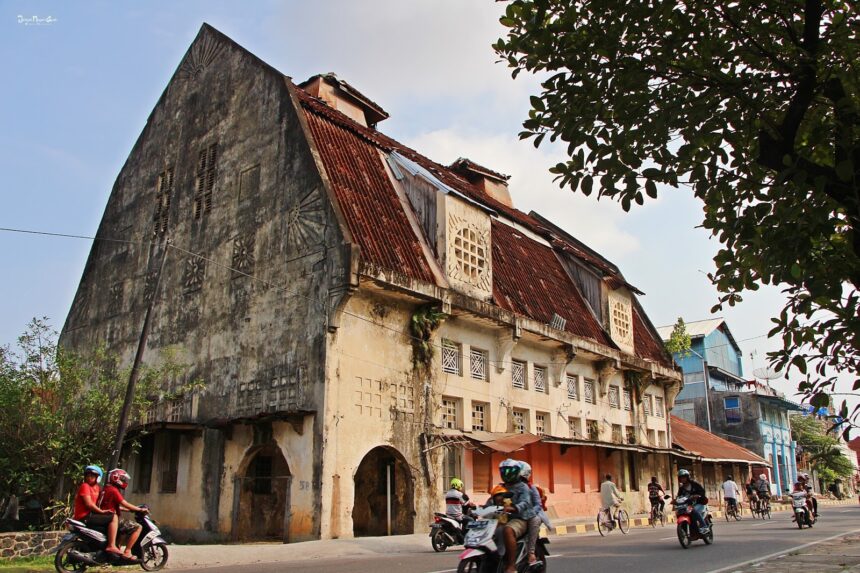 Weathered colonial-style building with a steep red-tiled roof on a busy street, motorbikes passing by in the foreground.