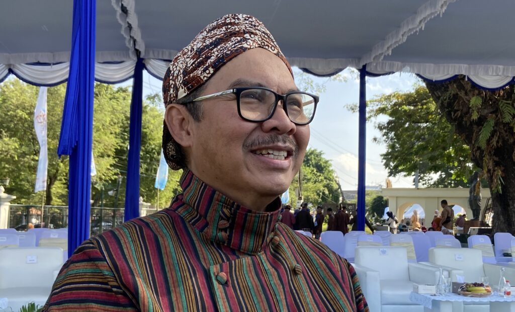 Close-up of a smiling man wearing glasses and a patterned headwrap at an outdoor event under a blue and white canopy.