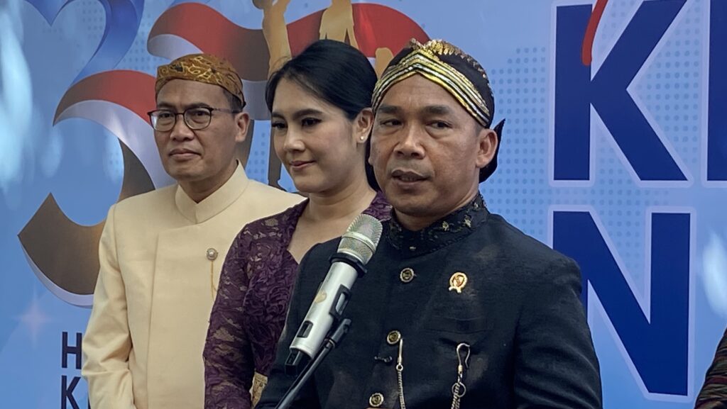 Three people in traditional Indonesian clothing at a press event; man in black speaks into a microphone while others stand beside him.