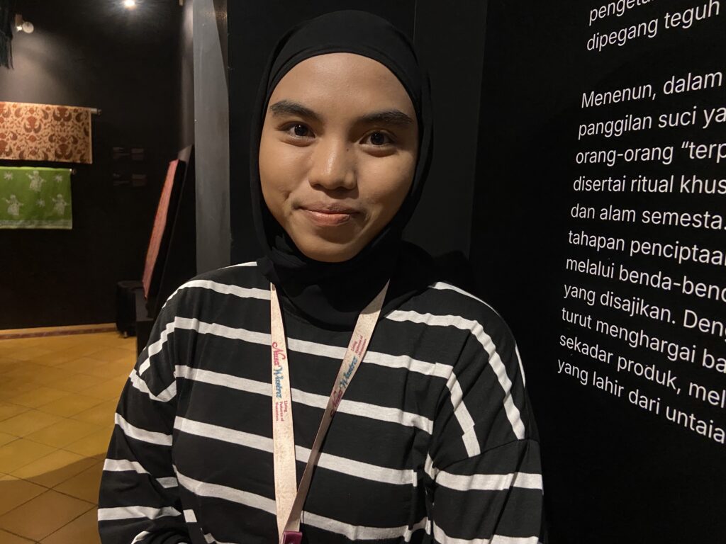 Close-up of a woman wearing a black hijab and striped top, smiling slightly, standing indoors beside a wall with white text.