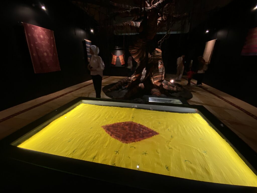Dimly lit museum exhibit with a large twisted tree sculpture surrounding a glowing yellow pool; visitors stand nearby to observe.