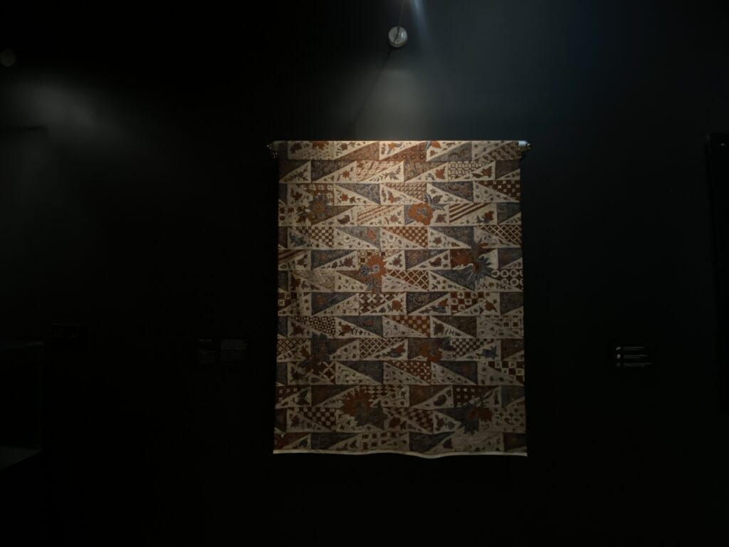 Patchwork quilt with geometric and floral triangle patches hangs on a dark gallery wall under a spotlight.