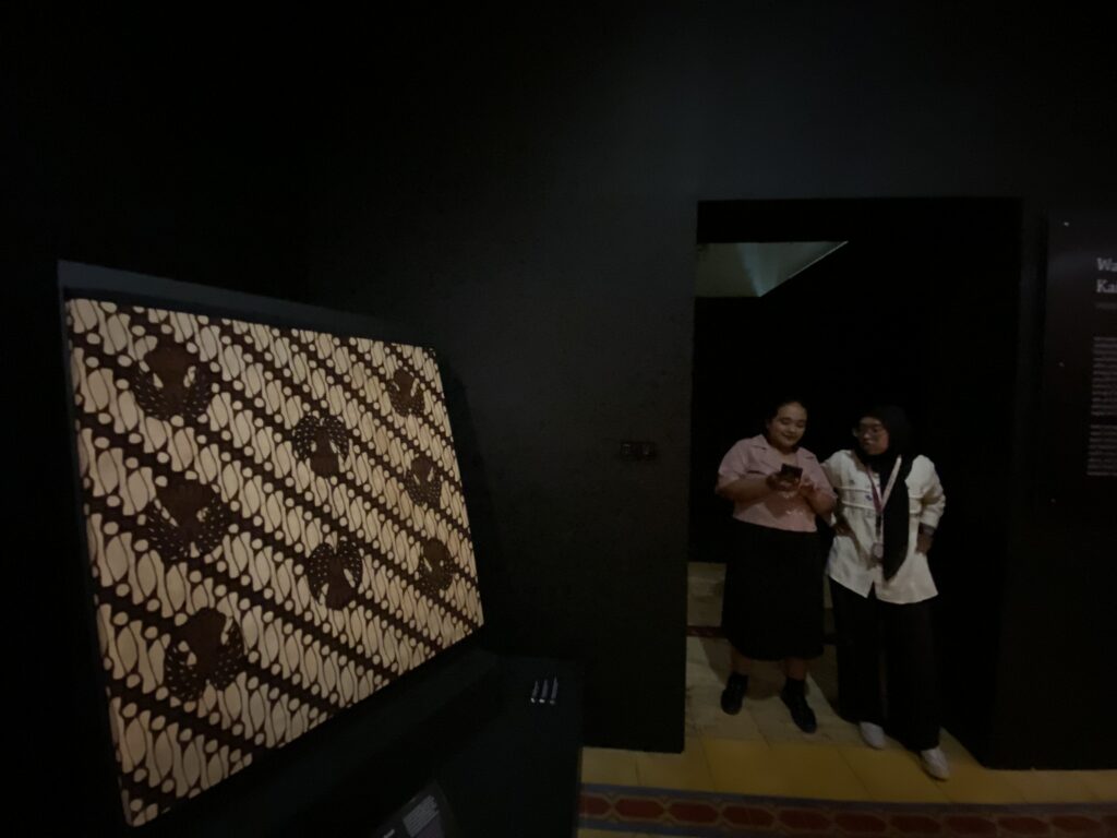 Dim gallery or museum space with a large black-and-white patterned textile on the left wall and two people in white coats standing near a dark doorway on the right.