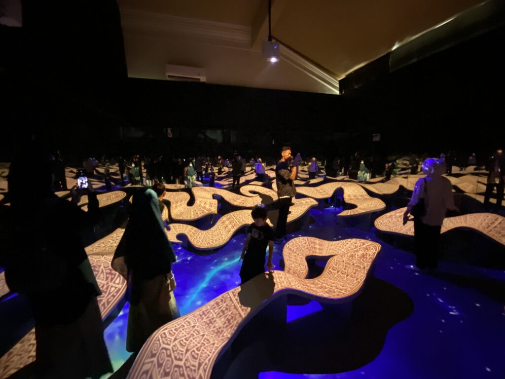 Dimly lit auditorium or theater with decorative patterned chairs and a standing crowd, blue stage lighting casting shadows on the floor.