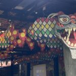 Dragon head sculpture with sharp teeth and orange eye, indoors among colorful hanging banners and lantern-like decorations.