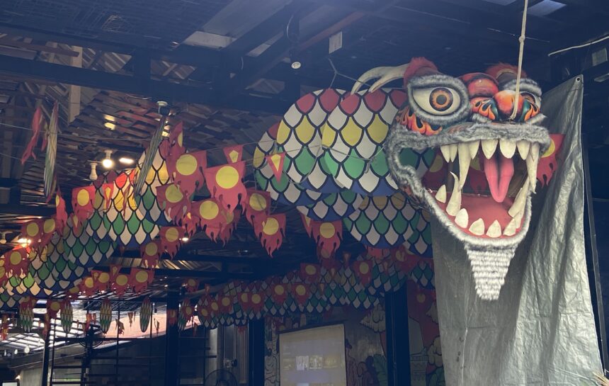 Dragon head sculpture with sharp teeth and orange eye, indoors among colorful hanging banners and lantern-like decorations.