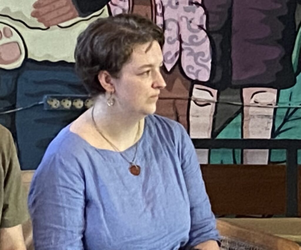 Woman with short dark hair wearing a blue top and a heart-shaped pendant, seated indoors with a mural in the background.