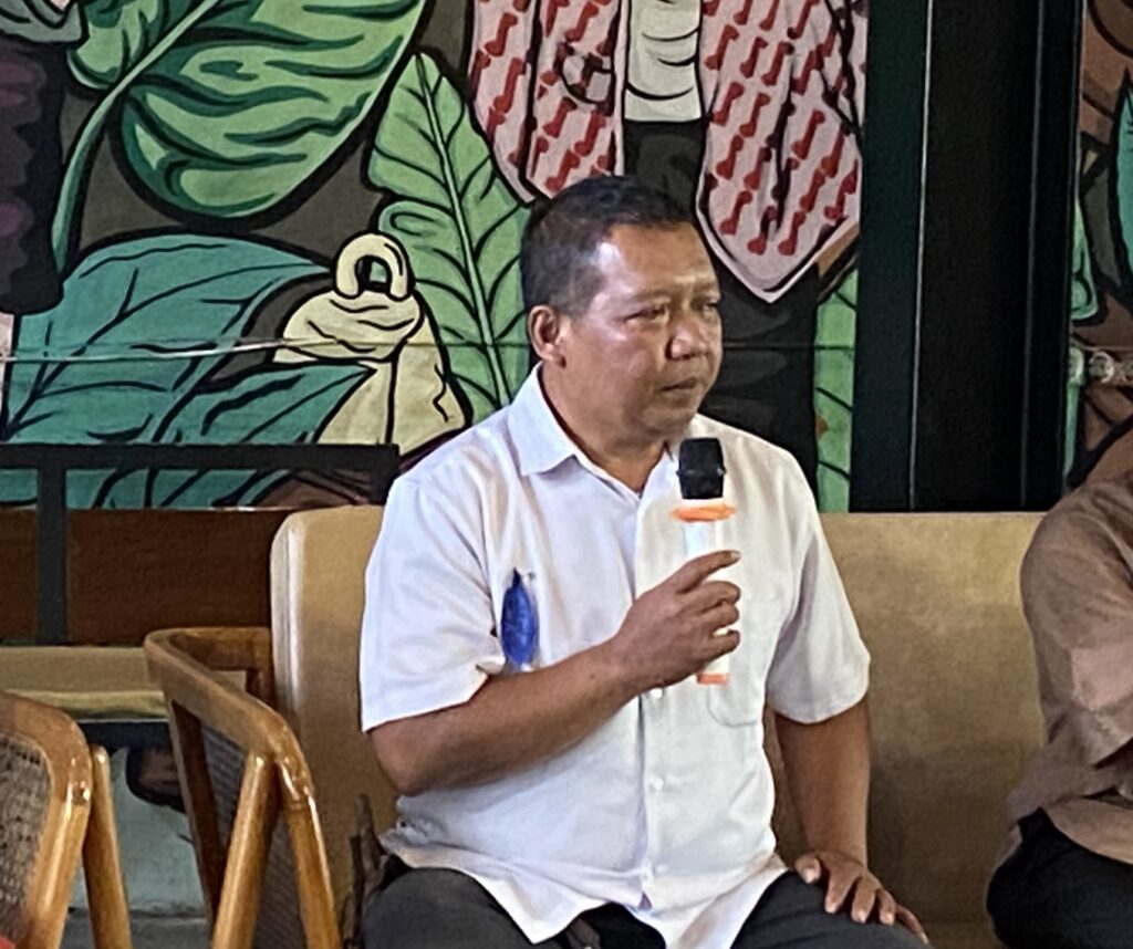 Man in a white shirt seated on a beige couch speaks into a handheld microphone, with a vibrant mural of green leaves behind him.