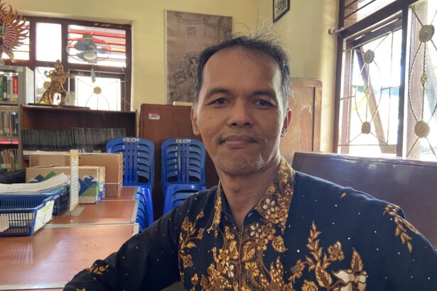 Close-up selfie of a man in a patterned batik shirt, smiling, with blue stacked chairs and shelves in a bright room behind him.