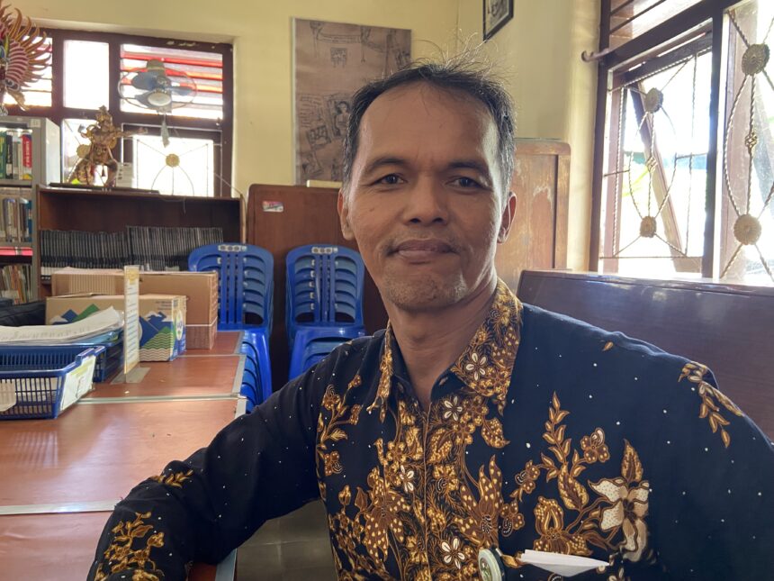 Close-up selfie of a man in a patterned batik shirt, smiling, with blue stacked chairs and shelves in a bright room behind him.