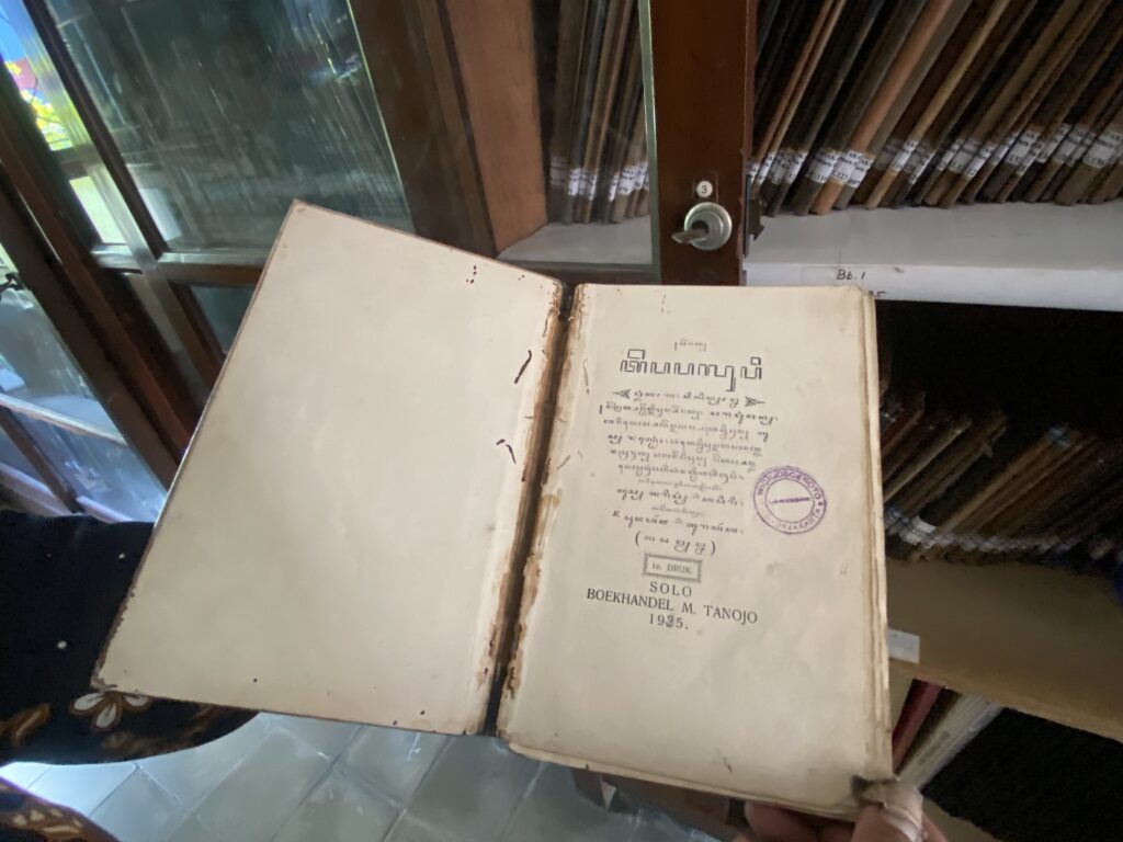 Open old book with a damaged spine resting on a surface, page showing printed text and a purple stamp, with wooden shelves of bound volumes in the background.