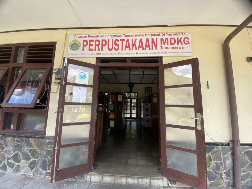 Open double doors to a library entrance with a large sign reading 'PERPUSTAKAAN MDKG Tamansiswa' above the doorway.