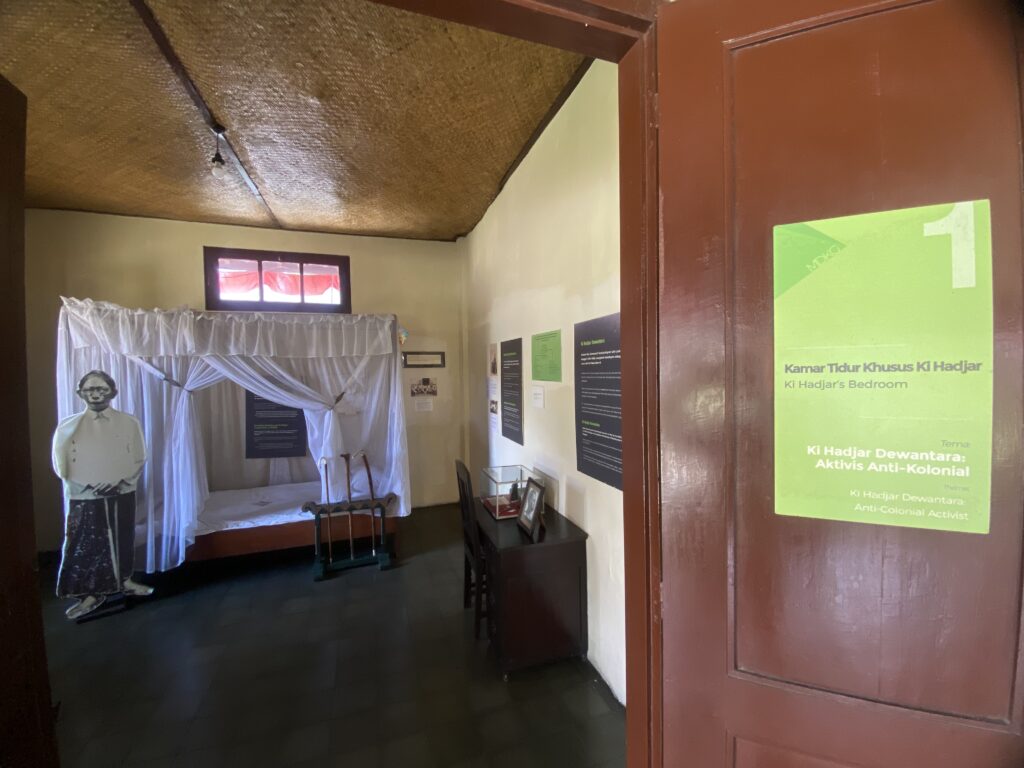 IMG_9651 - Mabur.co Museum room with a bed under a white canopy, a life-size man cutout, and a green exhibit sign on a brown door.