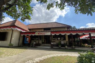 Front of a single-story building with red-tiled roof and red‑white bunting, under a blue sky with leafy tree branches above.