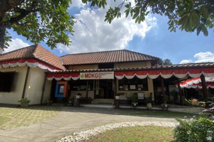 Front of a single-story building with red-tiled roof and red‑white bunting, under a blue sky with leafy tree branches above.