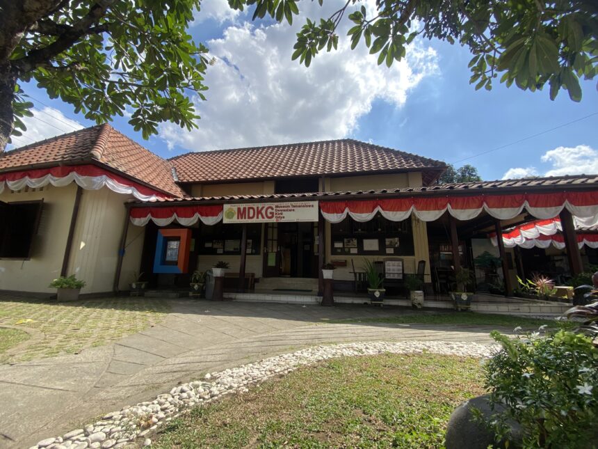 IMG_9654 - Mabur.co Front of a single-story building with red-tiled roof and red‑white bunting, under a blue sky with leafy tree branches above.