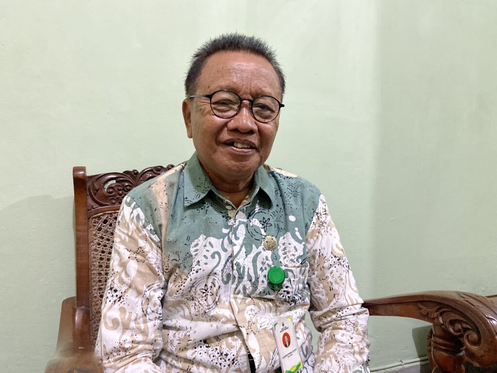 IMG_9662 - Mabur.co Older man with short grey hair and round glasses sits on a carved wooden chair, wearing a green patterned shirt and a lanyard badge.