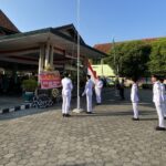 Line of students in white uniforms stand at attention in a flag-raising ceremony in a courtyard with an Indonesian flag nearby.