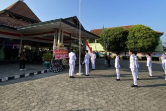 Line of students in white uniforms stand at attention in a flag-raising ceremony in a courtyard with an Indonesian flag nearby.