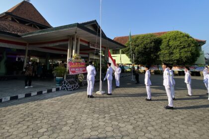 Line of students in white uniforms stand at attention in a flag-raising ceremony in a courtyard with an Indonesian flag nearby.