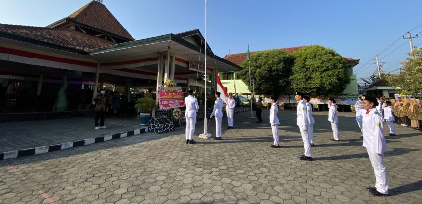 IMG_9724 - Mabur.co Line of students in white uniforms stand at attention in a flag-raising ceremony in a courtyard with an Indonesian flag nearby.