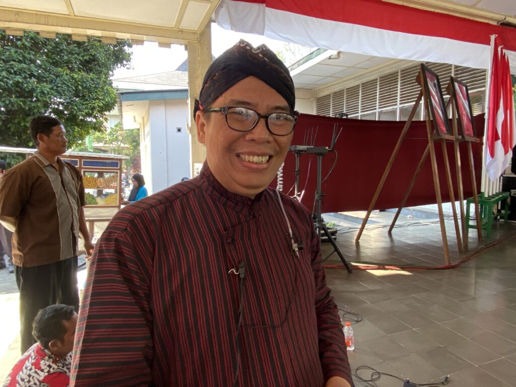 IMG_9738 - Mabur.co Smiling man wearing glasses and a black headwrap, in a red-striped shirt, at an outdoor event with red-and-white banners in the background.
