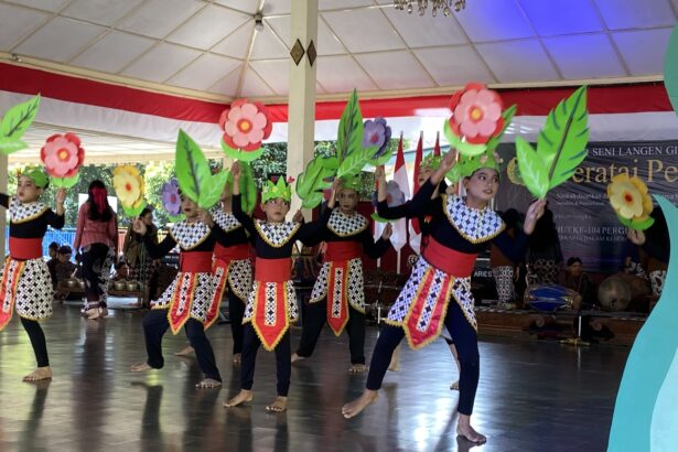 Group of young dancers in black-and-patterned costumes with red sashes performing on stage, holding large colorful paper flowers.