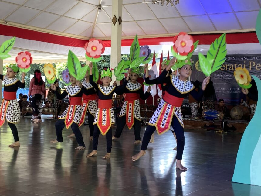 IMG_9753 - Mabur.co Group of young dancers in black-and-patterned costumes with red sashes performing on stage, holding large colorful paper flowers.
