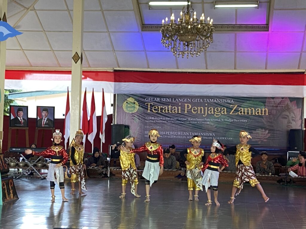 IMG_9756 - Mabur.co Dancers in ornate gold costumes perform on a stage with red-and-white flags and a large banner backdrop.