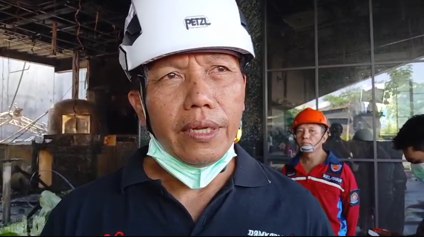 Man in a white Petzl hard hat speaking, with a green mask pulled under his chin at a construction site. -OR- A construction worker wearing a white helmet being interviewed, with others and debris in the background.