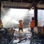 Firefighters in turnout gear spray water inside a charred building, with hoses and debris on the wet floor.