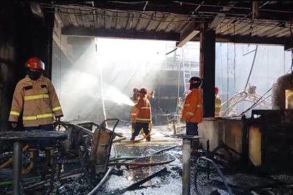Firefighters in turnout gear spray water inside a charred building, with hoses and debris on the wet floor.