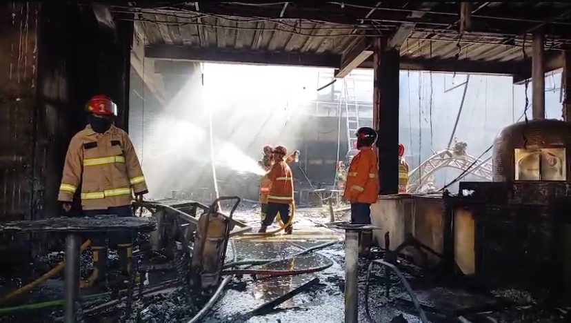 Firefighters in turnout gear spray water inside a charred building, with hoses and debris on the wet floor.