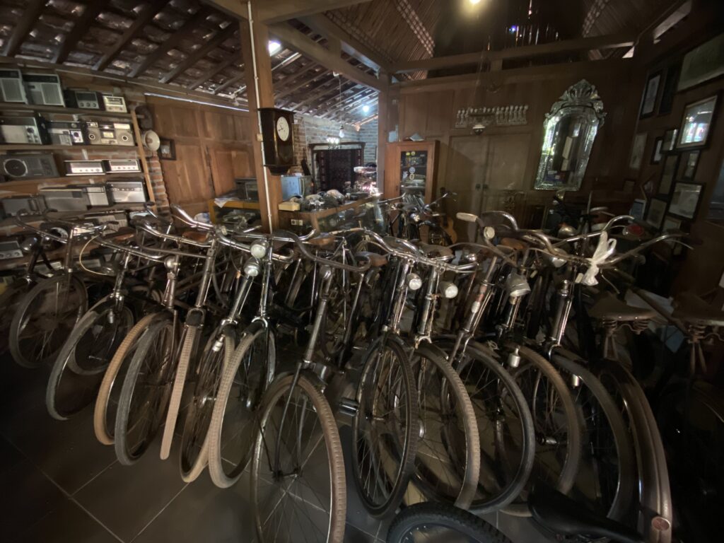 IMG_9808 - Mabur.co Cluttered storage room filled with numerous bicycles parked close together, against wooden walls with vintage radios and clocks on shelves nearby.