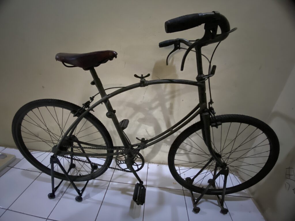 IMG_9838 - Mabur.co Indoor view of a black vintage bicycle leaning on a small wheeled repair stand against a beige wall, with saddle and curved handlebars visible inside a tiled room.