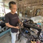 Man wearing a dark T-shirt stands in a cluttered workshop, holding bicycle parts over a workbench with tools and machinery nearby.